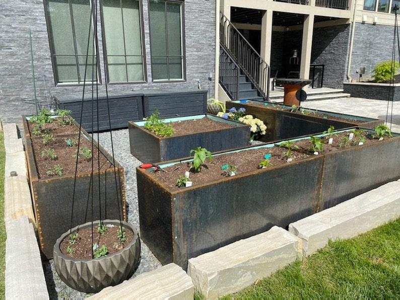 May include: A set of raised garden beds made of rusted metal, with a variety of plants growing in them. The beds are arranged in a U-shape and are surrounded by a stone border.