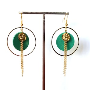 Op de afbeelding: Een paar groene en gouden oorbellen met een cirkelvormig ontwerp. De oorbellen hebben een groene cirkel met een gouden cirkel eromheen en een gouden kettingkwast die eronder hangt.