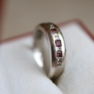 May include: A silver ring featuring square, deep red gemstones and smaller clear stones. The ring is presented in a white box with a red border, suitable for jewellery.