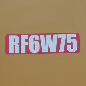 May include: A close-up of a yellow metal surface with a red rectangular sticker that says "RF6W75" in white letters.