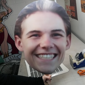 May include: A large cardboard cutout of a person's head with a smiling expression. The cutout is held by a hand.