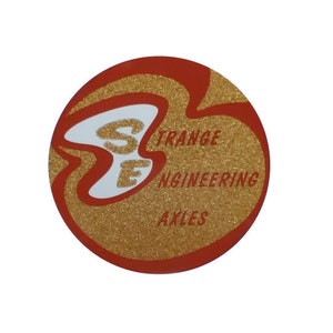 以下が含まれることがあります： 赤と金のグリッターデザインの丸いステッカー。ステッカーには、金色の文字で「STRANGE ENGINEERING AXLES」というテキストが記載されています。「S E」の文字は、白い様式化されたフォントです。