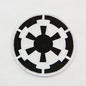 May include: Round black and white embroidered patch with a symmetrical design. The patch has a black centre with white geometric shapes radiating outwards, encircled by a white border. Suitable for clothing or accessories.