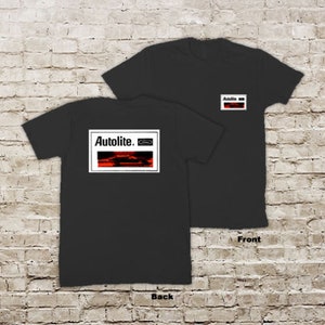May include: Black t-shirt with a white rectangle on the back that says "Autolite." Inside the rectangle is a red image of a car with the word "Ford" in a smaller white rectangle. The front of the shirt has a small white rectangle with the word "Autolite" in it.