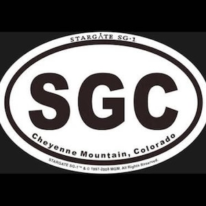 May include: White oval sticker with black text that reads "STARGATE SG-1" at the top, "SGC" in large letters in the center, and "Cheyenne Mountain, Colorado" at the bottom. The text "STARGATE SG-1™ & © 1997-2008 MGM All Rights Reserved." is printed in smaller letters at the bottom.