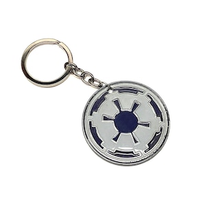 Puede incluir: Un llavero de metal plateado con un símbolo imperial blanco y azul de Star Wars. El símbolo es un círculo con una estrella en el centro.