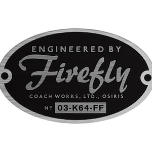 Puede incluir: Placa ovalada de metal negro y plateado con el texto "ENGINEERED BY Firefly COACH WORKS, LTD., OSIRIS" y el número "03-K64-FF". La placa tiene un borde de metal cepillado y dos pequeños agujeros.