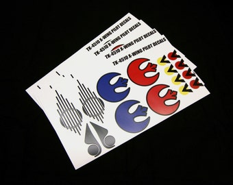Może przedstawiać: Stos białych naklejek z napisem "TK-4510 X-WING PILOT DECALS". Naklejki zawierają różne wzory, w tym niebieskie, czerwone i żółte symbole oraz czarno-szare kształty geometryczne.