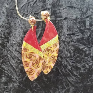 Könnte beinhalten: Ein Paar rote und goldene Blatt-Ohrringe mit einem floralen Muster. Die Ohrringe sind tropfenförmig und haben einen goldenen Akzent oben.