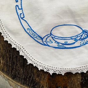 May include: White crocheted doily with a blue embroidered design of a teacup and saucer.