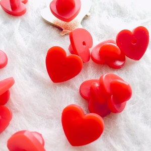 Peut inclure: Gros plan sur des punaises rouges en forme de cœur sur une surface blanche texturée. Les punaises ont un dessus arrondi et un dos plat avec une épingle. L'image met en valeur la couleur vive et la forme des punaises.