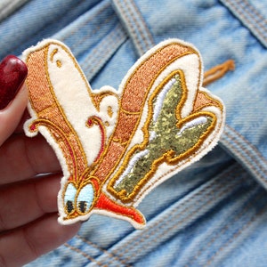 Peut inclure: Écusson brodé représentant un papillon de dessin animé. Le patch a un fond crème avec des détails brodés marron et or. Les ailes du papillon ont une section verte scintillante. Le patch est sur une veste en jean.