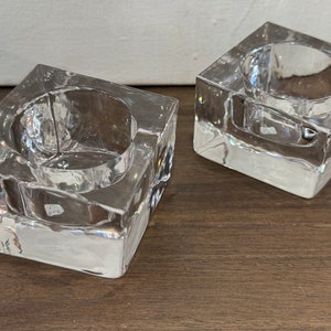 Puede incluir: Dos candelabros cuadrados de vidrio transparente. Cada uno tiene una hendidura circular para sostener una vela. El vidrio tiene una apariencia texturizada y facetada. Los candelabros están sobre una superficie de madera marrón oscuro.