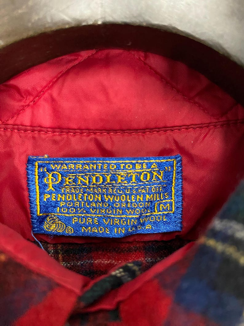 Puede incluir: Una etiqueta azul y blanca con el texto "WARRANTED TO BE A PENDLETON TRADE MARK REG US PAT OFF PENDLETON WOOLEN MILLS PORTLAND, OREGON 100% VIRGIN WOOL PURE VIRGIN WOOL MADE IN U.S.A." y una talla "M" en la esquina inferior derecha.