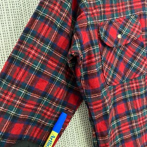 Puede incluir: Camisa de franela a cuadros rojos y verdes con un solo bolsillo en el pecho. La camisa tiene cuello abotonado y mangas largas.