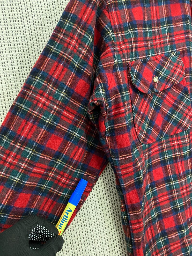 Puede incluir: Camisa de franela a cuadros rojos y verdes con un solo bolsillo en el pecho. La camisa tiene cuello abotonado y mangas largas.