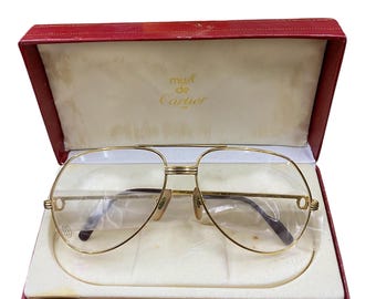 May include: Gold-tone aviator eyeglasses with clear lenses, presented in a red box. The glasses feature a double bridge and a subtle logo on the frame. The box is lined with a cream-colored material, and the text "must de Cartier" is visible.