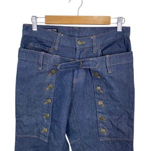 May include: Blue denim jeans with a unique design featuring a double waistband and a row of buttons down the side. The jeans are labeled "Final Home".