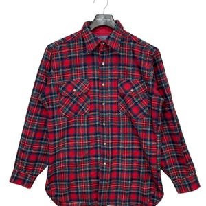 Puede incluir: Camisa de franela a cuadros rojos, verdes y negros con mangas largas y dos bolsillos en el pecho. La camisa est&aacute; colgada en una percha negra.