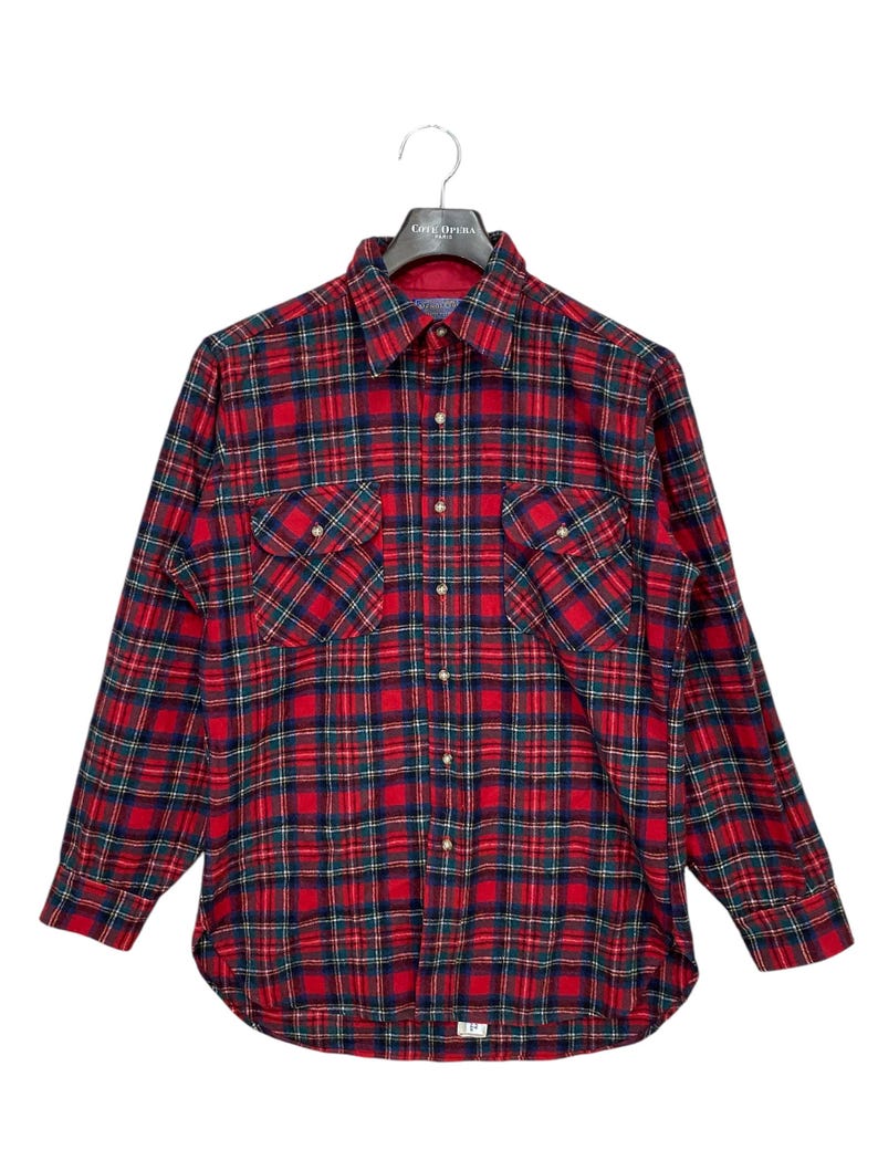 Puede incluir: Camisa de franela a cuadros rojos, verdes y negros con mangas largas y dos bolsillos en el pecho. La camisa est&aacute; colgada en una percha negra.