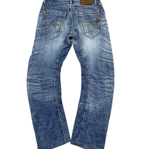 Può includere: Jeans blu in denim con lavaggio sbiadito e dettagli usurati. I jeans presentano il classico stile a cinque tasche, una toppa in pelle marrone sul retro e cuciture decorative sulle tasche posteriori. I jeans sono di un lavaggio medio con un leggero taglio a zampa.