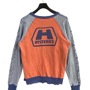 May include: A grey and orange sweatshirt with the word "HYSTERICS" printed on the back. The sleeves are grey with a blue design. The sweatshirt is hanging on a black hanger.