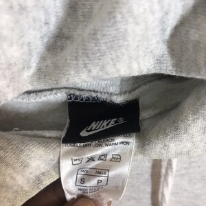 Peut inclure: Sweat &agrave; capuche gris Nike avec une &eacute;tiquette blanche indiquant "Nike" et des instructions de lavage. L'&eacute;tiquette indique &eacute;galement "Taille S, Taille P" et "Fabriqu&eacute; aux &Eacute;tats-Unis".