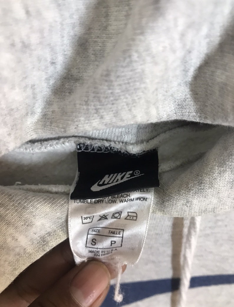 Peut inclure: Sweat &agrave; capuche gris Nike avec une &eacute;tiquette blanche indiquant "Nike" et des instructions de lavage. L'&eacute;tiquette indique &eacute;galement "Taille S, Taille P" et "Fabriqu&eacute; aux &Eacute;tats-Unis".