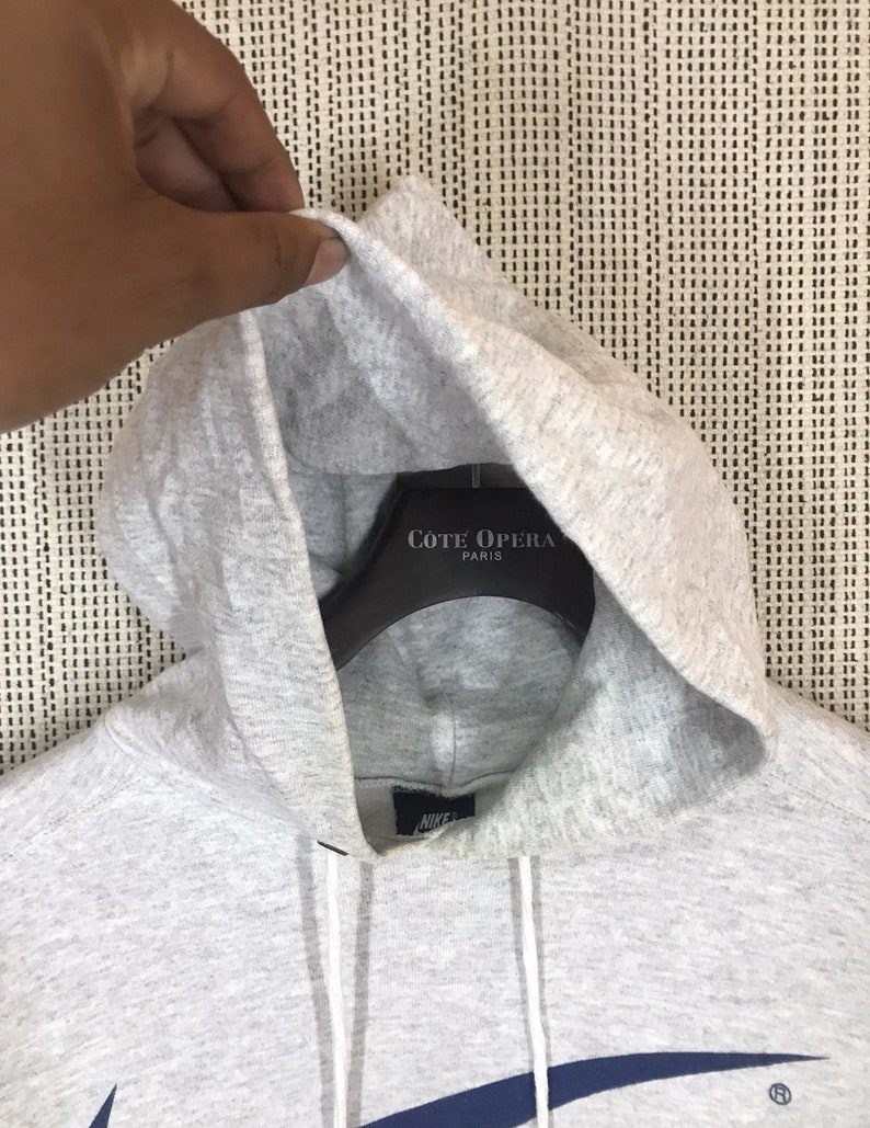 Peut inclure: Sweat-shirt &agrave; capuche gris avec un logo Nike swoosh bleu marine et le mot "Nike" en lettres blanches sur le devant. La capuche est doubl&eacute;e de tissu gris et a un cordon de serrage blanc. Le sweat-shirt a une &eacute;tiquette blanche avec le texte "C&ocirc;te Opera Paris" dessus.