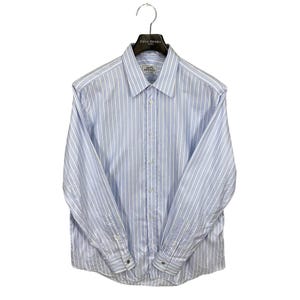 Puede incluir: Camisa a rayas azules y blancas con mangas largas, colgada de una percha negra. La camisa tiene cuello y puños abotonados. La etiqueta de la camisa dice "Hermès".