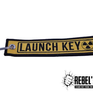 Op de afbeelding: Een gele en zwarte rechthoekige sleutelhanger met de tekst "LAUNCH KEY" en een stralingssymbool. De tag heeft een metalen sleutelring bevestigd. Het merklogo "REBEL'55" staat in de rechter benedenhoek.