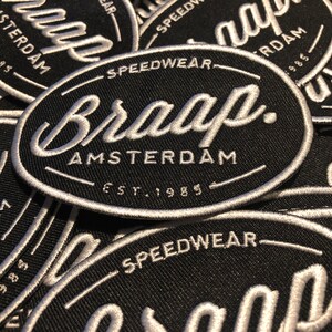 Op de afbeelding: Zwarte ovale patch met witte geborduurde tekst. De patch leest "Braap Amsterdam" met "Speedwear" erboven en "Est. 1985" eronder. Meerdere patches zijn zichtbaar, wat een gestructureerd effect creëert.
