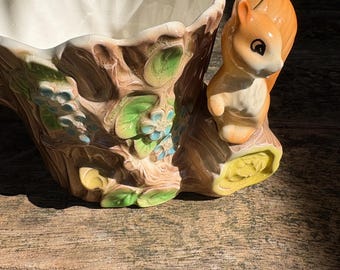 May include: A ceramic planter shaped like a tree trunk with a squirrel figurine. The planter is cream-colored inside and brown on the outside, with green and blue floral accents. The squirrel is orange and white.