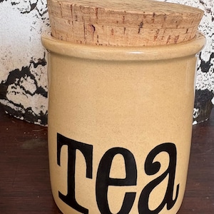 Puede incluir: Un tarro de té de cerámica de color beige con tapa de corcho. El tarro tiene la palabra "Tea" escrita en letras negras.