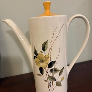 May include: A white ceramic coffee pot with a yellow lid and a floral design featuring a yellow flower with green leaves and brown stems.