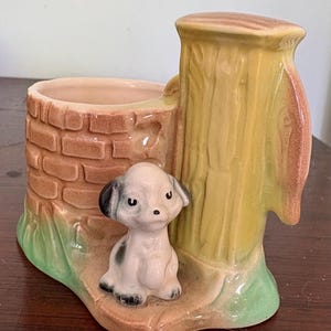 May include: A ceramic planter with a brick well design and a seated puppy figurine. The planter has a light brown interior, a yellow tree trunk, and a green base. The puppy is white with black spots and ears.