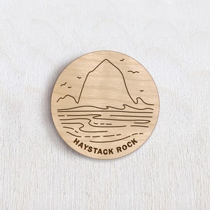 May include: A round, wooden coaster with a laser-etched design of Haystack Rock. The design includes a mountain, waves, birds, and the text "HAYSTACK ROCK" in a simple font. The coaster is made of light-colored wood.