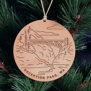 May include: A wooden Christmas ornament with a laser-engraved design of a bridge over a river with mountains in the background. The ornament is brown and has the text "DECEPTION PASS, WA" on the bottom.
