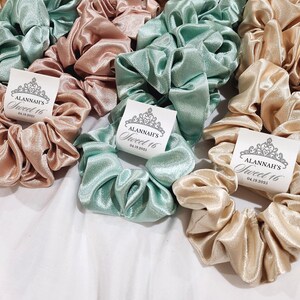 May include: A collection of satin scrunchies in light green and beige with personalized tags that say "Alannah's Sweet 16" and the date "04.19.2023".