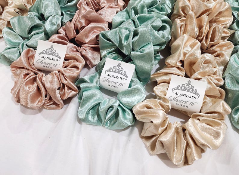 May include: A collection of satin scrunchies in light green and beige with personalized tags that say "Alannah's Sweet 16" and the date "04.19.2023".
