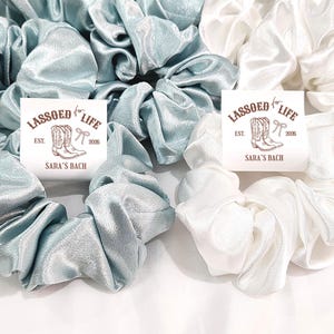 May include: Scrunchies in shades of white and teal blue are displayed. Each scrunchie is made of a shiny, silky fabric. The image includes cards with the text "LASSOED LIFE" and "SARA'S BACH" with a cowboy boot graphic.