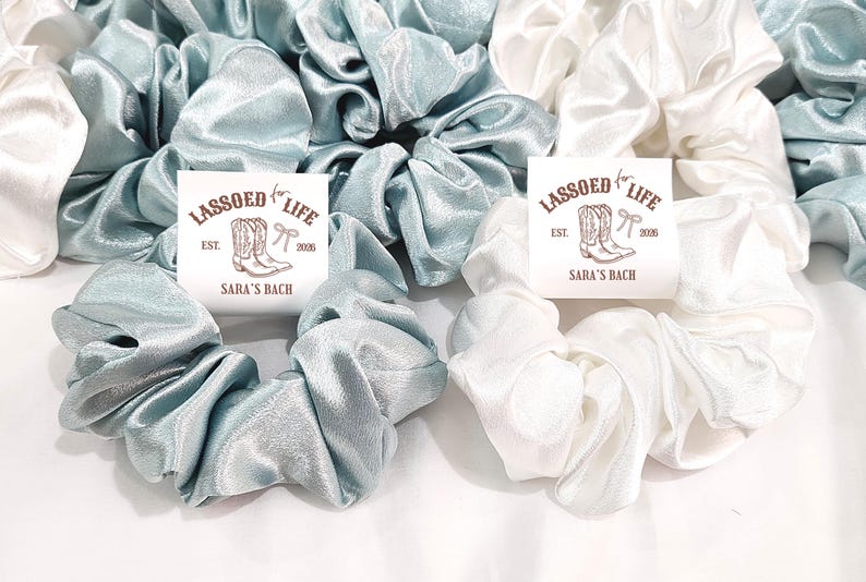 May include: Scrunchies in shades of white and teal blue are displayed. Each scrunchie is made of a shiny, silky fabric. The image includes cards with the text "LASSOED LIFE" and "SARA'S BACH" with a cowboy boot graphic.