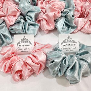 May include: A set of light blue and pink satin scrunchies with a white card attached to each. The card has a crown design and the text "Alannah's Sweet 16 04.19.2023".