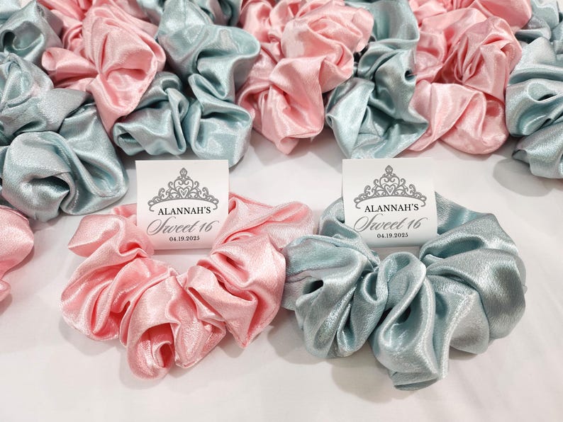 May include: A set of light blue and pink satin scrunchies with a white card attached to each. The card has a crown design and the text "Alannah's Sweet 16 04.19.2023".