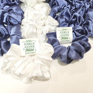 May include: A collection of satin scrunchies in white and a muted blue. The scrunchies are gathered together, with some displaying a small tag that reads "GIRLS GONE Mild" and "Jenn's Bach" in green text.