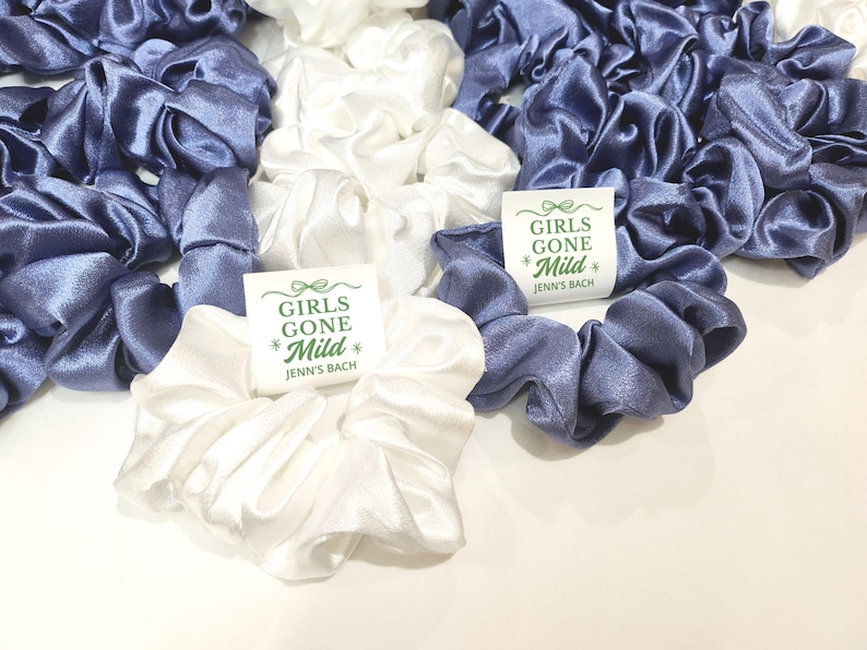 May include: A collection of satin scrunchies in white and a muted blue. The scrunchies are gathered together, with some displaying a small tag that reads "GIRLS GONE Mild" and "Jenn's Bach" in green text.