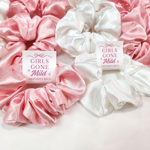 May include: A selection of satin scrunchies in pink and white. The scrunchies are gathered and voluminous. Some have a small white tag with the text "GIRLS GONE Mild" and "HEATHER'S BACH" in pink. These are likely hair accessories.