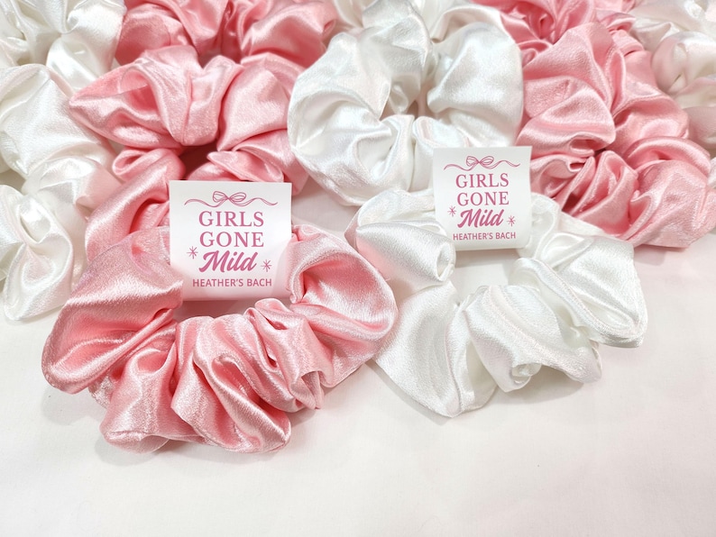 May include: A selection of satin scrunchies in pink and white. The scrunchies are gathered and voluminous. Some have a small white tag with the text "GIRLS GONE Mild" and "HEATHER'S BACH" in pink. These are likely hair accessories.