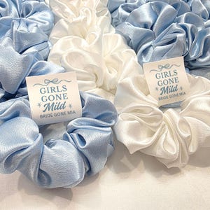 May include: A selection of light blue and white satin scrunchies. Each scrunchie has a small tag that reads "GIRLS GONE Mild" and "Bride Gone Mia". The scrunchies are grouped together, giving a soft, textured look.