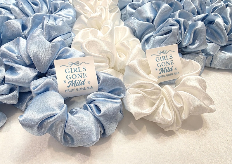 May include: A selection of light blue and white satin scrunchies. Each scrunchie has a small tag that reads "GIRLS GONE Mild" and "Bride Gone Mia". The scrunchies are grouped together, giving a soft, textured look.